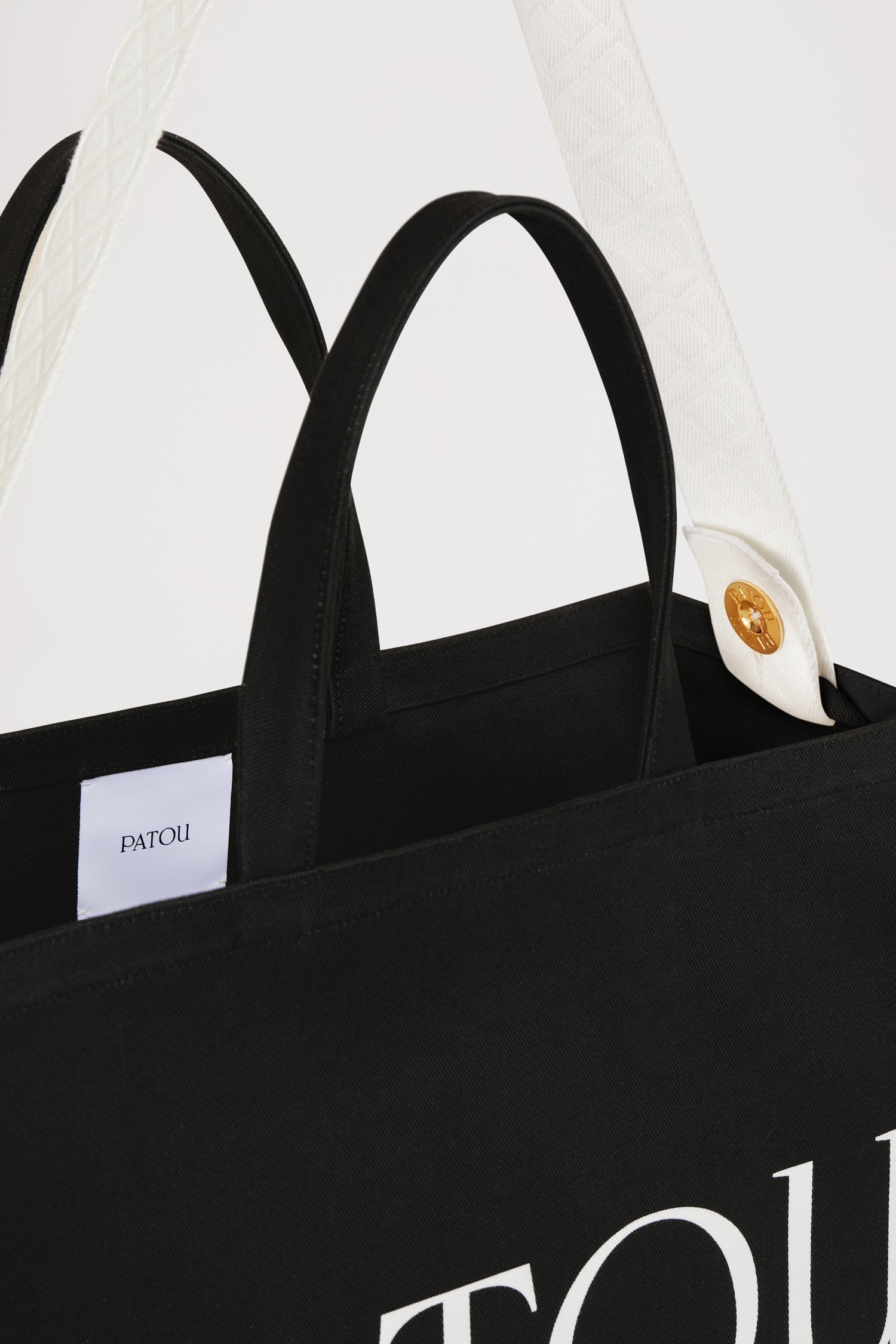 Patou - Patou canvas tote in organic cotton - Black - Carry your Patou everywhere with you. Black organic cotton tote. Patou lettering logo and JP monogram in white. White embossed shoulder strap with gold buttons. Two top handles. Can be carried by hand or on the shoulder.