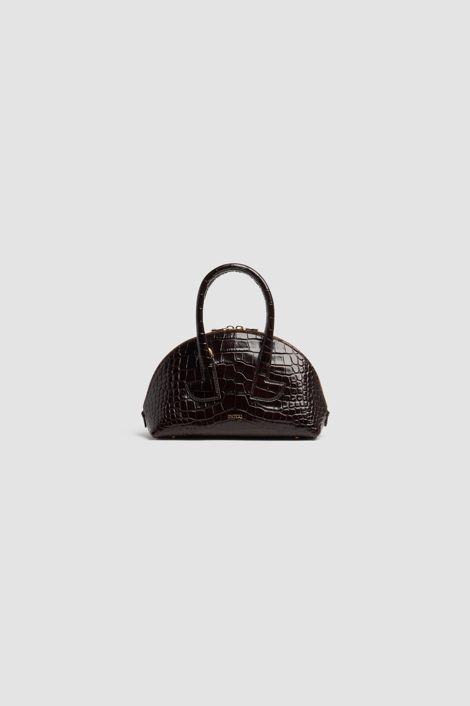 Patou - Borsa Le Patou Bowling in pelle di coccodrillo goffrata - Smoked Ebony - Borsa in pelle di coccodrillo goffrata Smoked Ebony. Forma bowling. Doppio manico con dettagli geometrici. Tracolla rimovibile. Chiusura con zip oro a doppio cursore. Logo Patou oro goffrato sul davanti. Piedini in metallo oro.