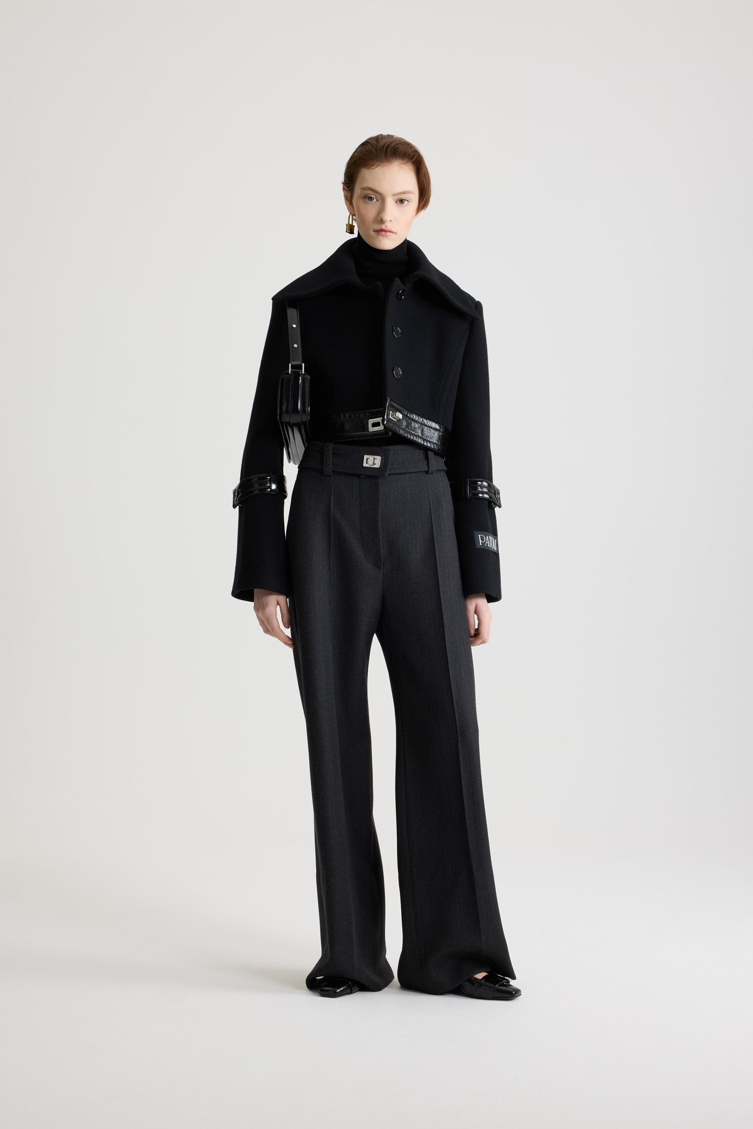 Patou - サステナブル ウールブレンド クロップド ロックジャケット - Black - ブラックのサステナブル ウール混紡ジャケット。丈の短いシルエット。シングルブレストスタイル。ワイドカラー。隠しボタン留め。シルバーカラーのロック留め付きのパテントレザー製ウエストバンド。袖にパテントレザー製のボタン付きタブ。左袖にPatouロゴラベル。裏地付き。