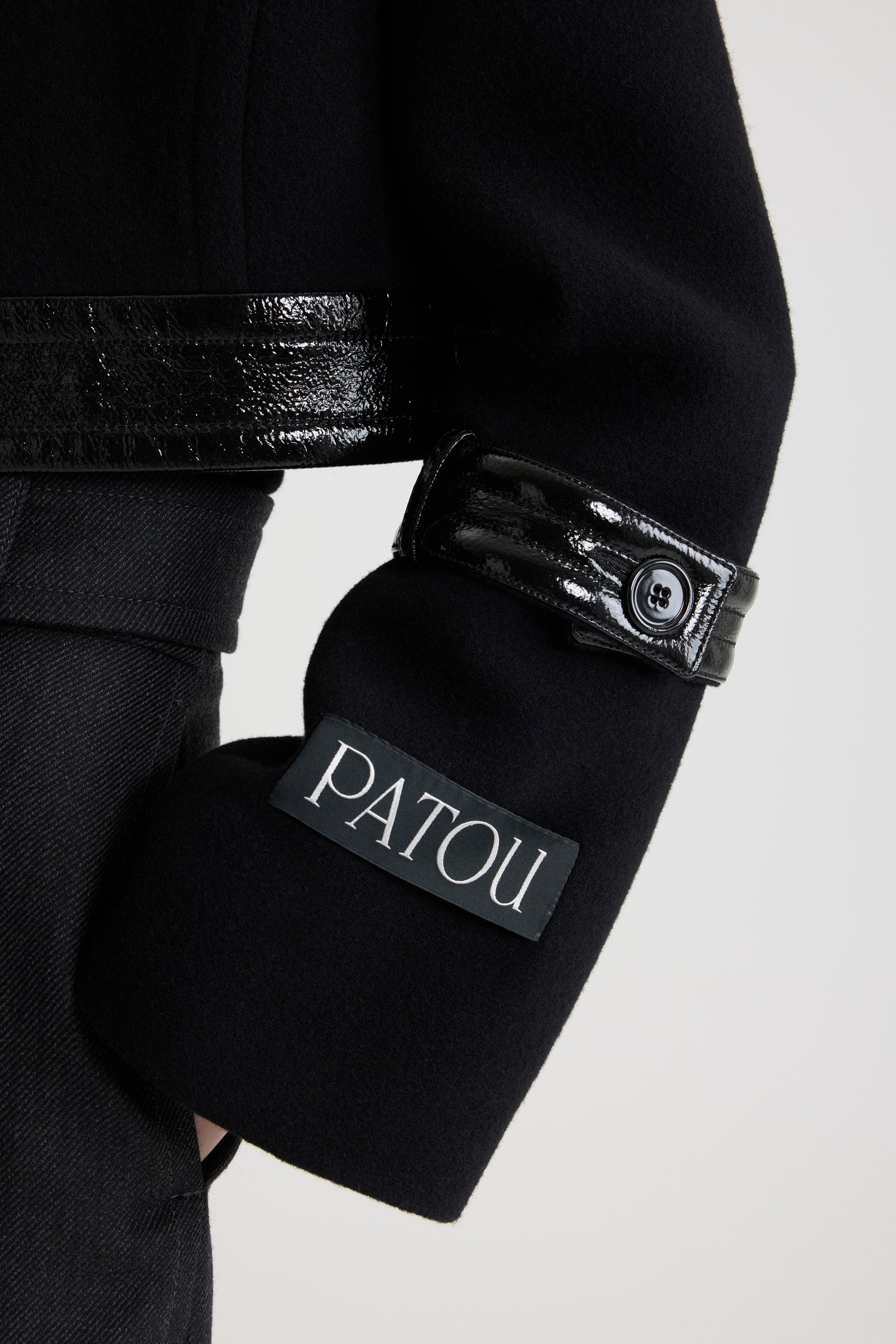 Patou - サステナブル ウールブレンド クロップド ロックジャケット - Black - ブラックのサステナブル ウール混紡ジャケット。丈の短いシルエット。シングルブレストスタイル。ワイドカラー。隠しボタン留め。シルバーカラーのロック留め付きのパテントレザー製ウエストバンド。袖にパテントレザー製のボタン付きタブ。左袖にPatouロゴラベル。裏地付き。