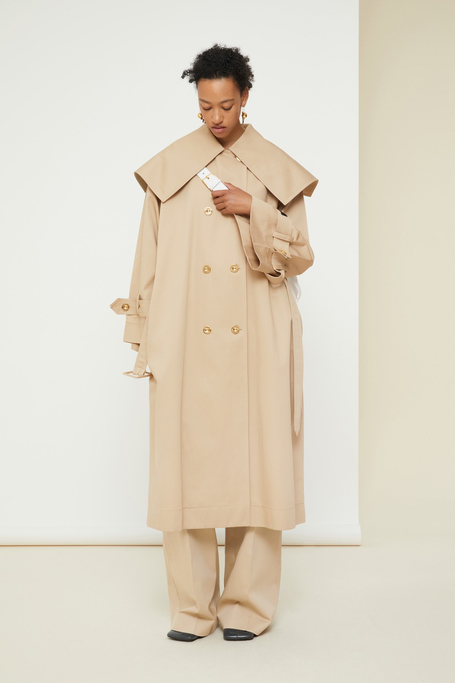 Patou - Charakteristischer Trenchcoat aus Bio-Baumwolle - Sand - Ein ausgestellter Trenchcoat mit französischem Flair. Charakteristischer Patou Trenchcoat aus sandbeiger Bio-Baumwoll-Gabardine. Doppelreihig mit leichter A-Linie. Übergroßer Kragen und Sturmklappe. Goldfarbene Knöpfe mit Patou Logo-Gravur. Abnehmbarer Taillengürtel. Bindebänder an den Manschetten mit schwarzem gestickten Patou Etikett. Zwei Seitentaschen. Vollständig gefüttert.