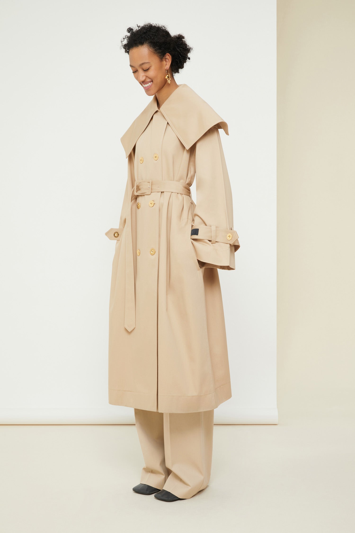 Patou - Charakteristischer Trenchcoat aus Bio-Baumwolle - Sand - Ein ausgestellter Trenchcoat mit französischem Flair. Charakteristischer Patou Trenchcoat aus sandbeiger Bio-Baumwoll-Gabardine. Doppelreihig mit leichter A-Linie. Übergroßer Kragen und Sturmklappe. Goldfarbene Knöpfe mit Patou Logo-Gravur. Abnehmbarer Taillengürtel. Bindebänder an den Manschetten mit schwarzem gestickten Patou Etikett. Zwei Seitentaschen. Vollständig gefüttert.
