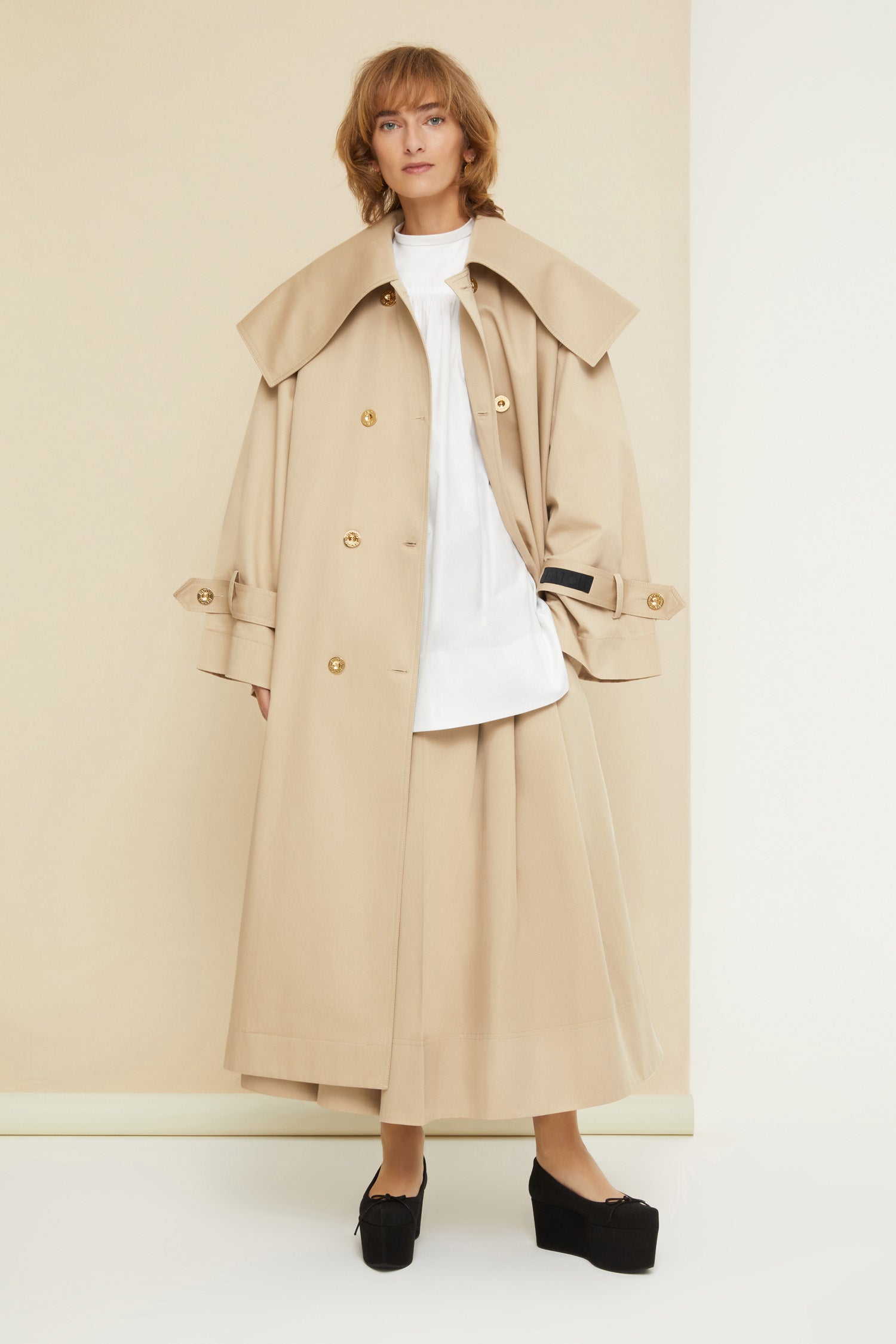 Patou - Charakteristischer Trenchcoat aus Bio-Baumwolle - Sand - Ein ausgestellter Trenchcoat mit französischem Flair. Charakteristischer Patou Trenchcoat aus sandbeiger Bio-Baumwoll-Gabardine. Doppelreihig mit leichter A-Linie. Übergroßer Kragen und Sturmklappe. Goldfarbene Knöpfe mit Patou Logo-Gravur. Abnehmbarer Taillengürtel. Bindebänder an den Manschetten mit schwarzem gestickten Patou Etikett. Zwei Seitentaschen. Vollständig gefüttert.