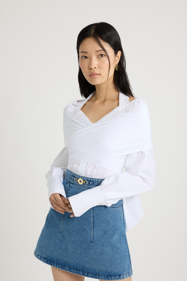 Patou - オーガニック コットンポプリン ラップブラウス - White - ホワイトのオーガニックコットンポプリン製ブラウス。バスト周りに巻きつけて背面で結ぶ、取り外し可能なトップレイヤーデザイン。伸縮性のあるショルダーとグログランホルターネックストラップを配したワイドなネックライン。隠しボタン留め。袖口にボタン付きの長めのスリーブ。前面にJPモノグラム刺繍。