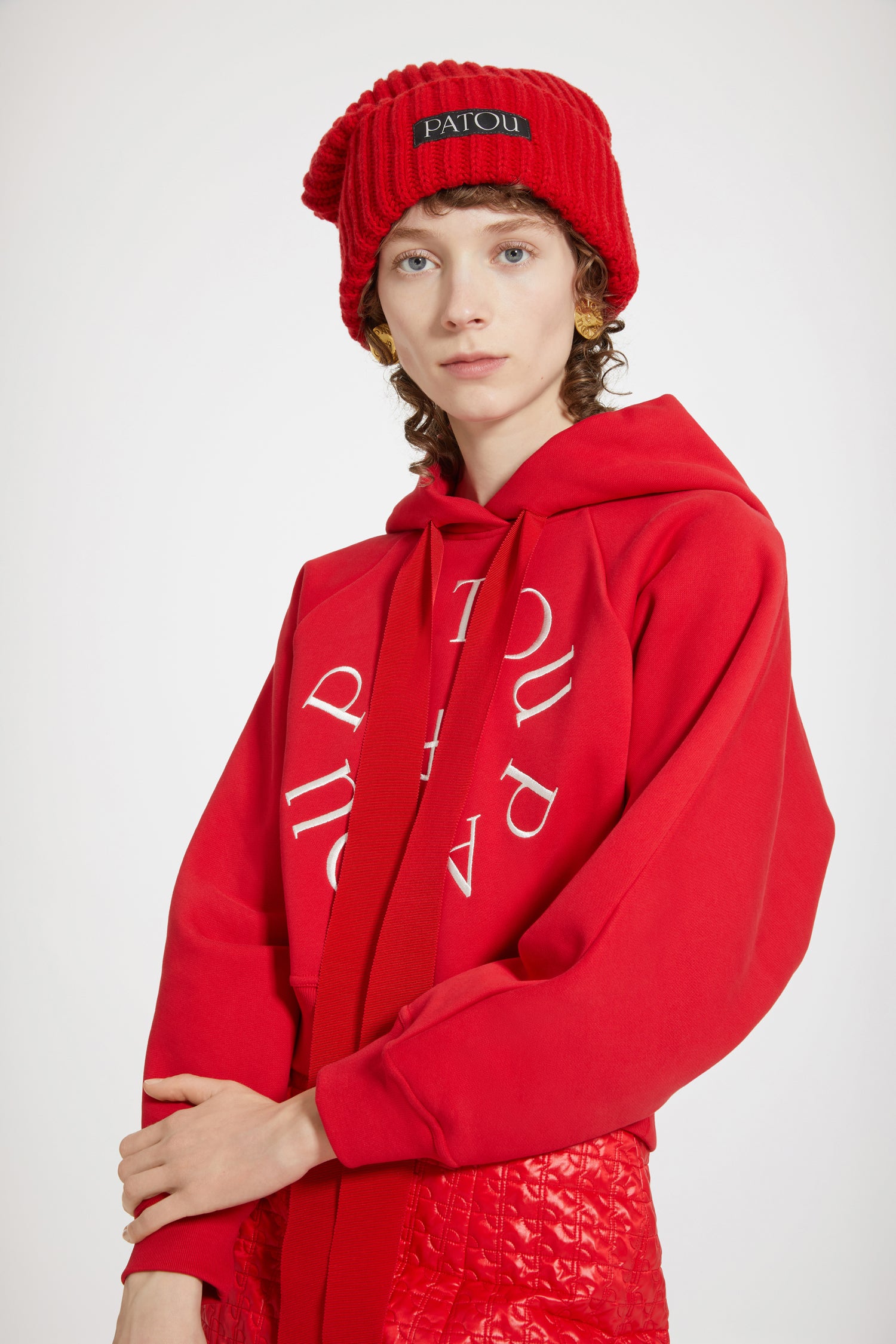 Patou - Felpa corta con logo medaglione in cotone bio - Racing Red - Variazione di stagione sulla felpa Patou.Felpa in cotone bio rosso. Modello corto. Collo con cappuccio e lacci in grosgrain. Logo Patou medaglione ricamato sul davanti. Maniche raglan. Polsini e vita a costine.