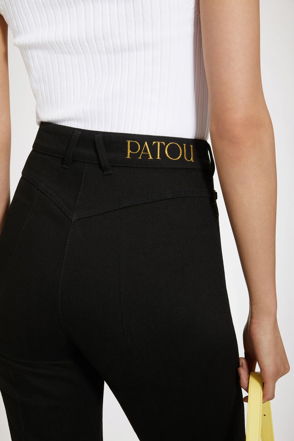 Patou - オーガニックコットンデニム製 フレアーパンツ - Black - ジャン・パトゥのアートが昇華された新しいパトゥデニム。ブラックカラーのオーガニックコットンデニム製パンツ。フレアーカット。ミッドライズ。フロントにゴールドトーンのパトゥ刻印入りボタン。ベルトループ付き。ウエストバンドにパトゥの刺しゅうロゴ。フロントとバックにダーツ使いとセンター位置の折り目。フロントにゴールドカラーのJP刻印入りオーナメントをあしらったポケット2つ。バックにブランドを象徴するカーブシーム。The ornament can be removed.